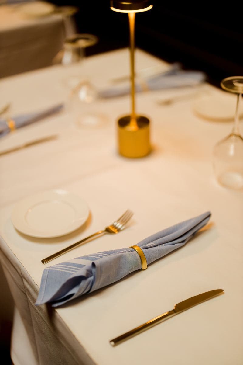 A chambray napkin in a brass ring beside gold flatware on white linen